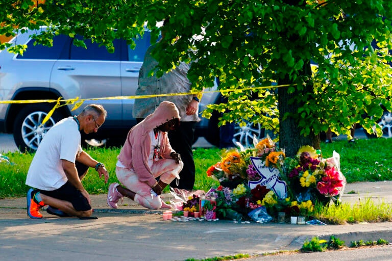 Two people kneel on the ground in front of a memorial made of flowers.