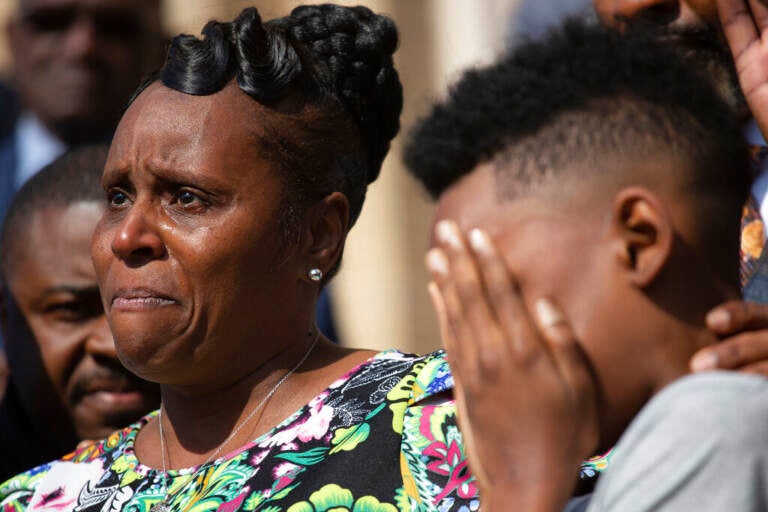 A young boy hides his face in his hands, while a woman in the background holds back tears.