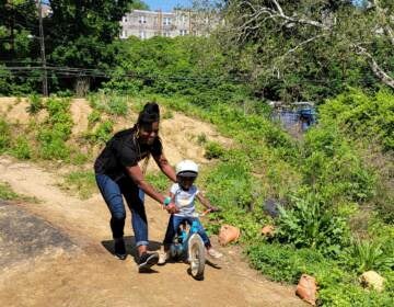 A woman supports a child on a small bike on a pumptrack.