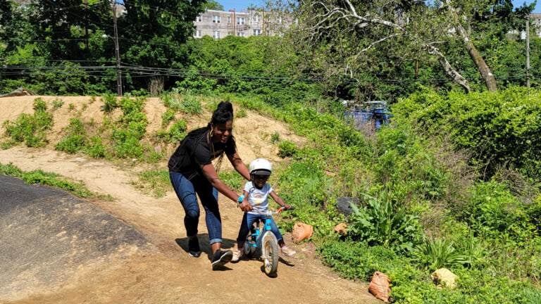 A woman supports a child on a small bike on a pumptrack.