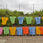 Rows of T-shirts bearing the name of Philadelphians lost to gun violence are on display with a fence and bushes in the background.