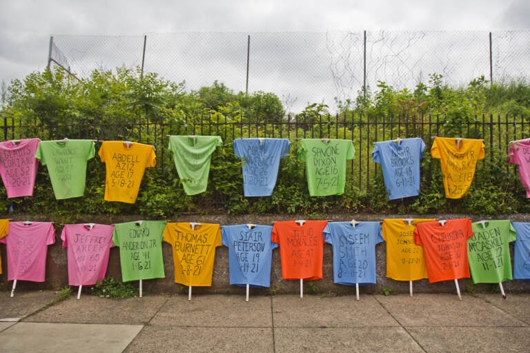 Rows of T-shirts bearing the name of Philadelphians lost to gun violence are on display with a fence and bushes in the background.