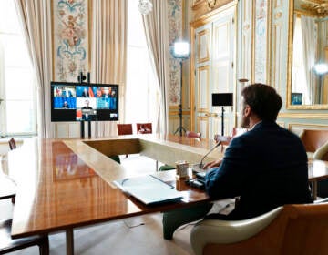 French President Emmanuel Macron is seen from behind, seated at a table, participating in a video conference with leaders of the other G7 countries.