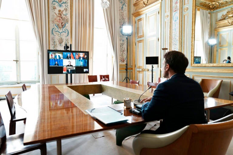 French President Emmanuel Macron is seen from behind, seated at a table, participating in a video conference with leaders of the other G7 countries.