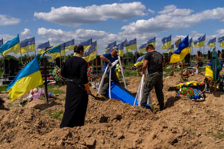 People gather around a grave in a dirt field with numerous Ukrainian flags flying.