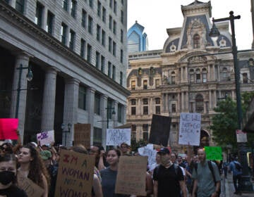 A large crowd of people demonstrates, holding signs, with City Hall visible in the background.