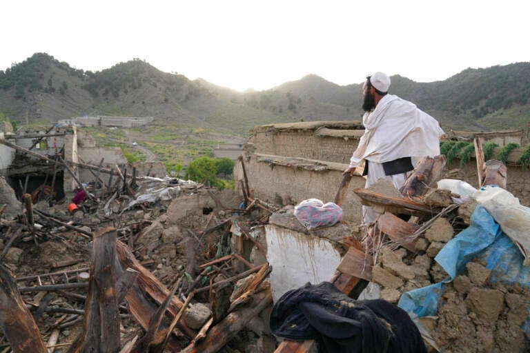 A man stands looking out over destroyed buildings, with mountains in the distance.