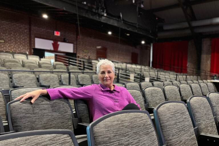 Carmen Khan sits in the middle of the audience seats of a theater, with her arm stretched out over the chair next to her.