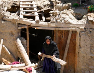 A woman carries a piece of wood as she walks outside through the entrance of a damaged house.