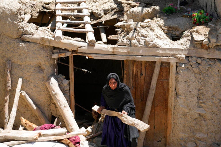A woman carries a piece of wood as she walks outside through the entrance of a damaged house.