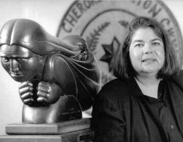 Portrait of Wilma Mankiller in black and white with a statue behind her.