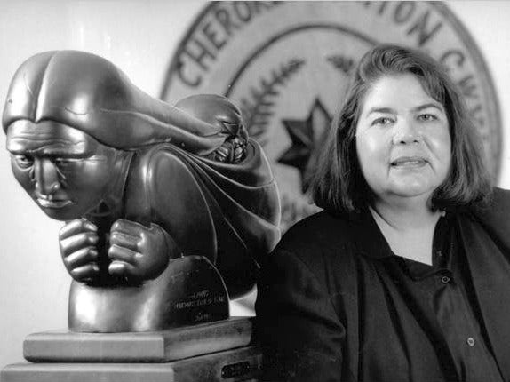 Portrait of Wilma Mankiller in black and white with a statue behind her.