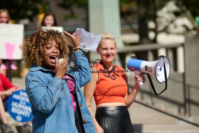 Rep. Minor-Brown talks into a microphone, as someone else behind her holds a megaphone in the air.