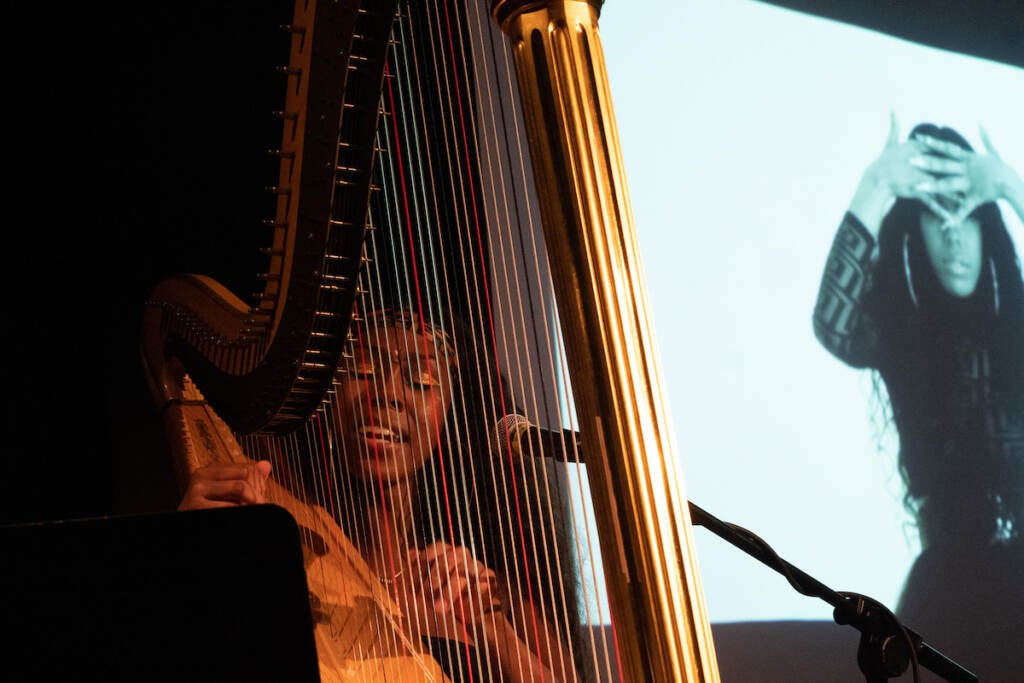 A woman plays the harp with a black and white video or photo shown in the background on a large screen.