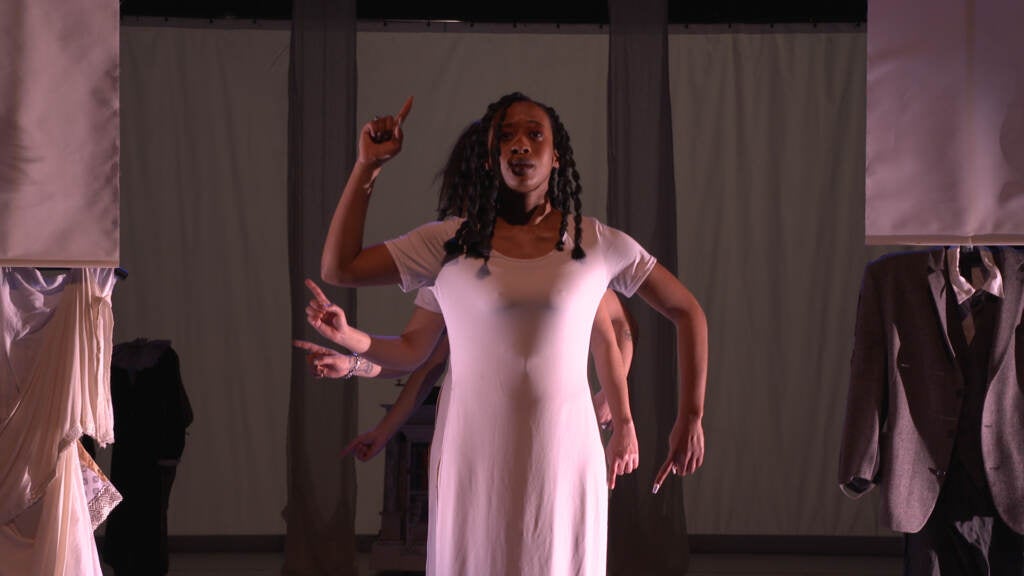 A person wearing a white dress stands on stage in dramatic lighting. 