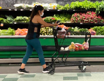 A shopper pushes their shopping cart in a grocery store, with fresh produce visible in the background.