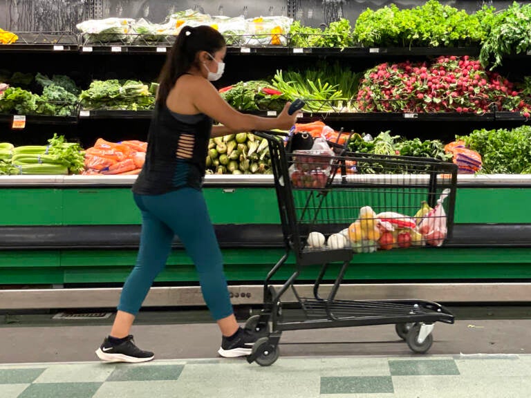 A shopper pushes their shopping cart in a grocery store, with fresh produce visible in the background.