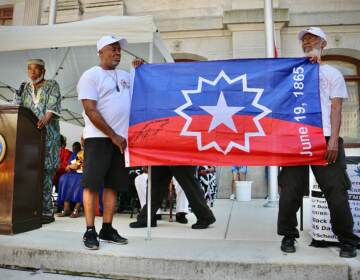 Members of the Black Male Community Council hold up the official flag of Juneteenth while Ben Haith (left) the creator of that flag describes what inspired him. Haith was the guest of honor at the flag raising ceremony at Philadelphia City Hall. (Emma Lee/WHYY)