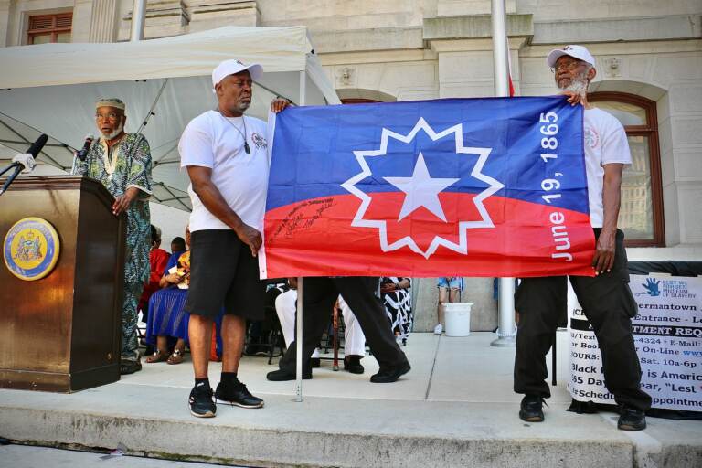 Members of the Black Male Community Council hold up the official flag of Juneteenth while Ben Haith (left) the creator of that flag describes what inspired him. Haith was the guest of honor at the flag raising ceremony at Philadelphia City Hall. (Emma Lee/WHYY)