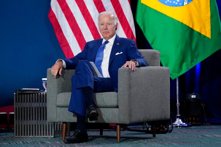 President Biden sits in a chair, with the U.S. and Brazilian flags behind him, at the Summit of the Americas.