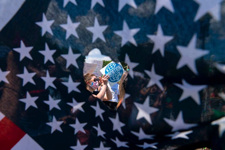 Abortion-rights activists are seen through a hole in an American flag as they protest outside the Supreme Court