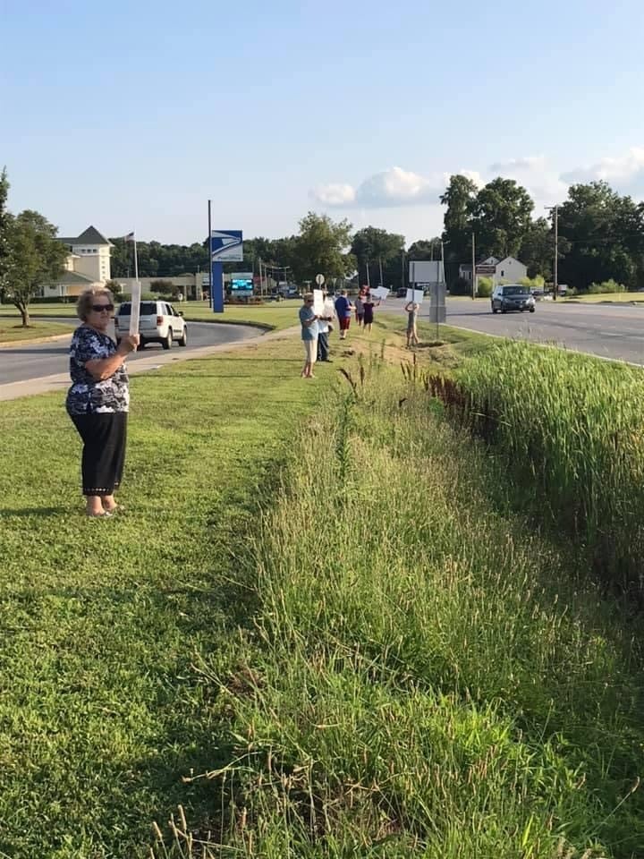 People stand in green grass on the side of a road holding signs.