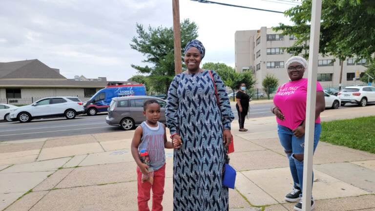 A mother stands holding her child's hand and smiling, with the sidewalk and street in the background.