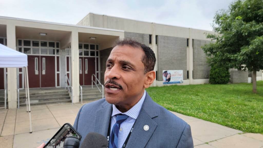 Close-up of Superintendent Watlington speaking into a microphone, with Alain Locke School visible in the background.
