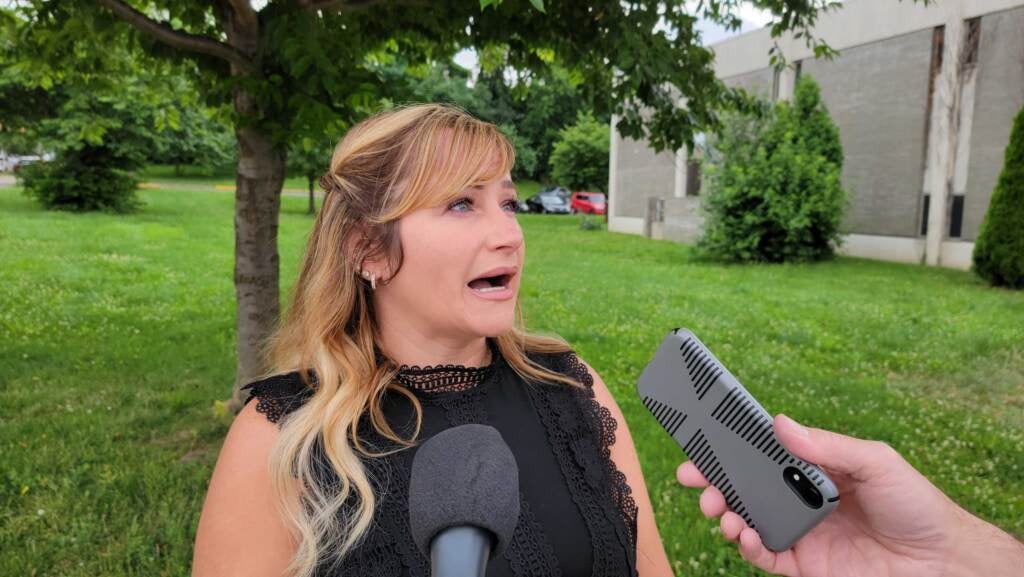 Waleska Maldonado speaks into several microphones, with grass, trees and a school building visible behind her.
