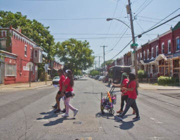 People cross a street in West Philadelphia, with brick rowhomes lining either side of the street.
