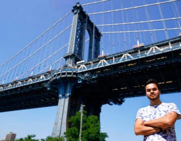A person stands with their arms crossed, with a bridge and blue sky visible in the background.
