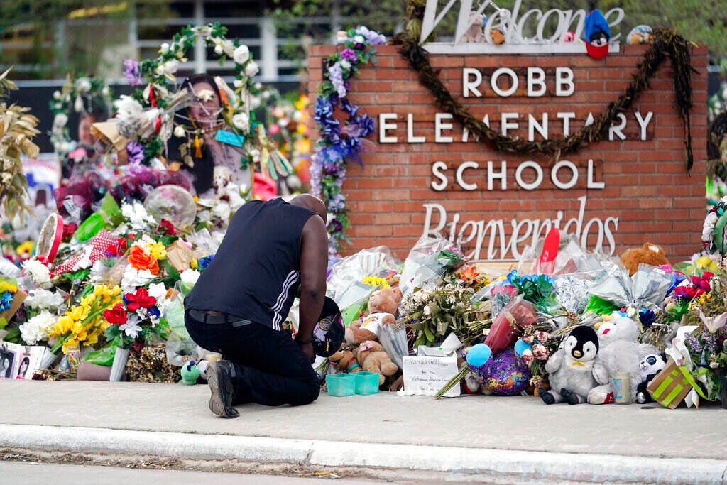 A man kneels in front of a memorial surrounding the sign that reads, "Robb Elementary School, Bienvenidos".