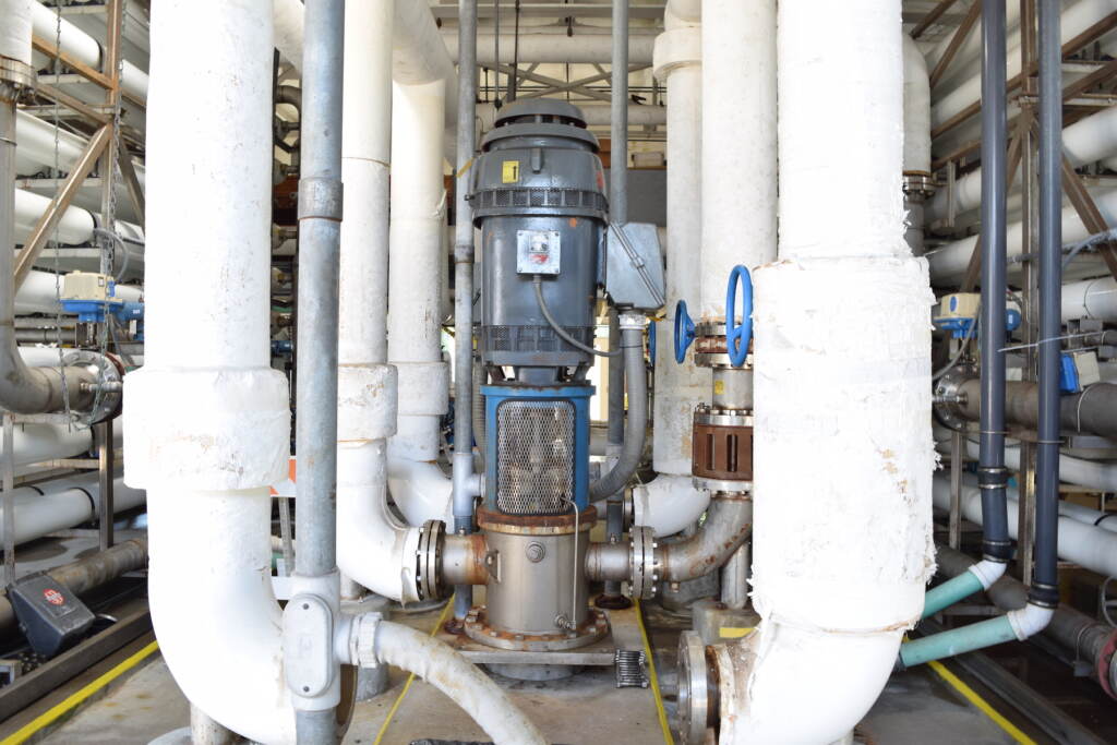 Several vertical white pipes surround a grey pump in the center of a room inside the Cape May Water Works building. 