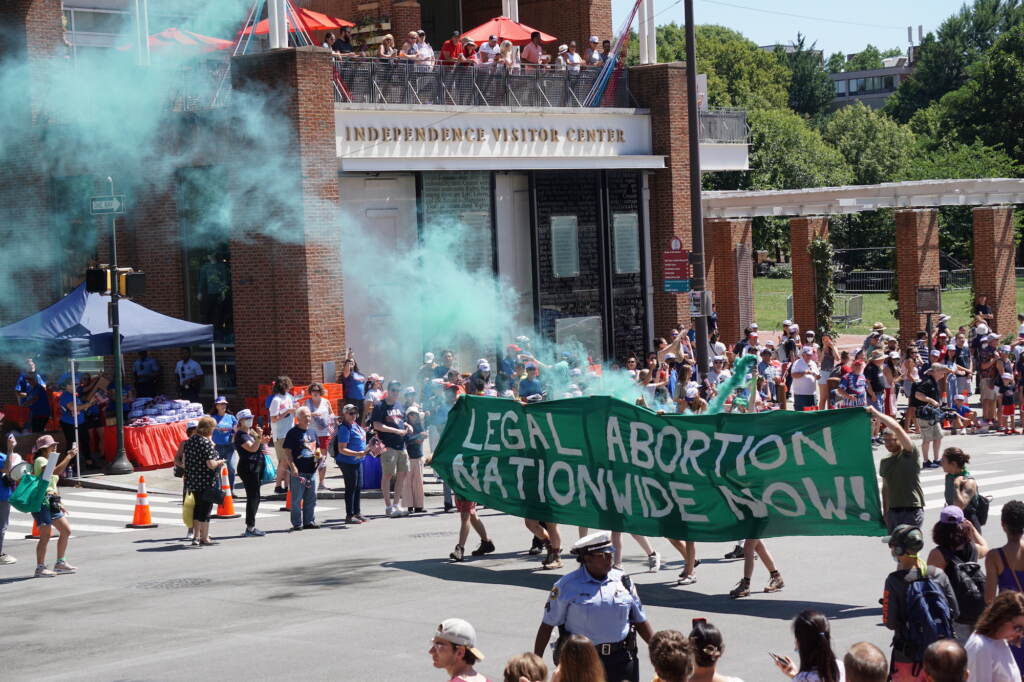 Protesters walk down the street amid green smoke with a sign that reads, "Legal Abortion Nationwide Now!".