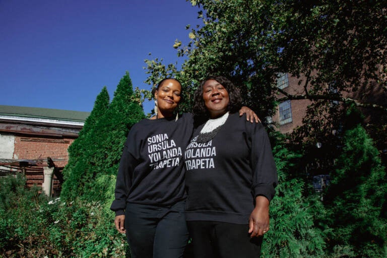 Two women stand together, facing the camera and smiling, with trees and a brick building visible in the background.