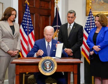 President Biden sits at a desk, signing an order, as Vice President Kamala Harris and others stand and look on.