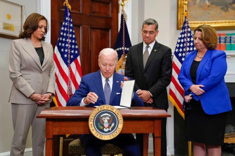 President Biden sits at a desk, signing an order, as Vice President Kamala Harris and others stand and look on.