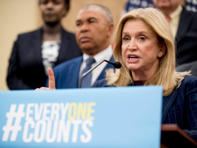 Carolyn Maloney Someone speaks at a podium with a sign reading,