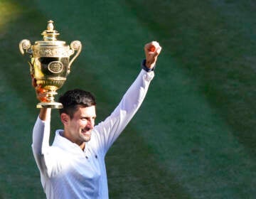 Djokovic holds the Wimbledon trophy up in the air with one hand as he raises his arms.