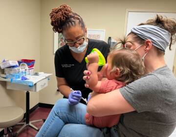 A doctor leans forward as she vaccinates a young child, who is crying as she is held by her mother.