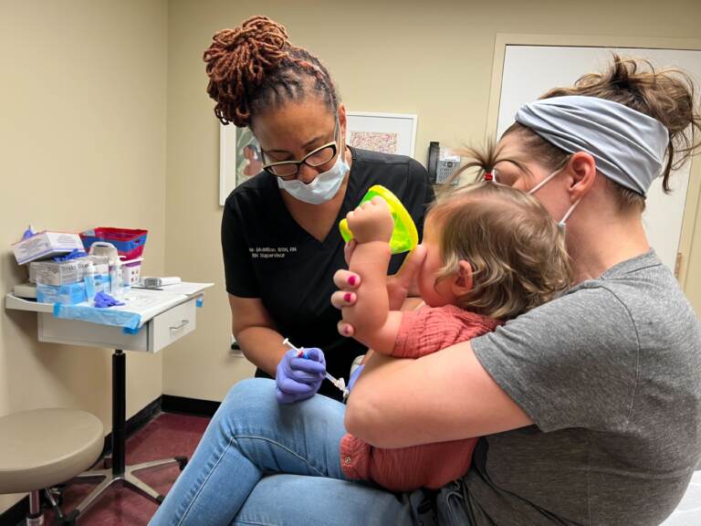 A doctor leans forward as she vaccinates a young child, who is crying as she is held by her mother.