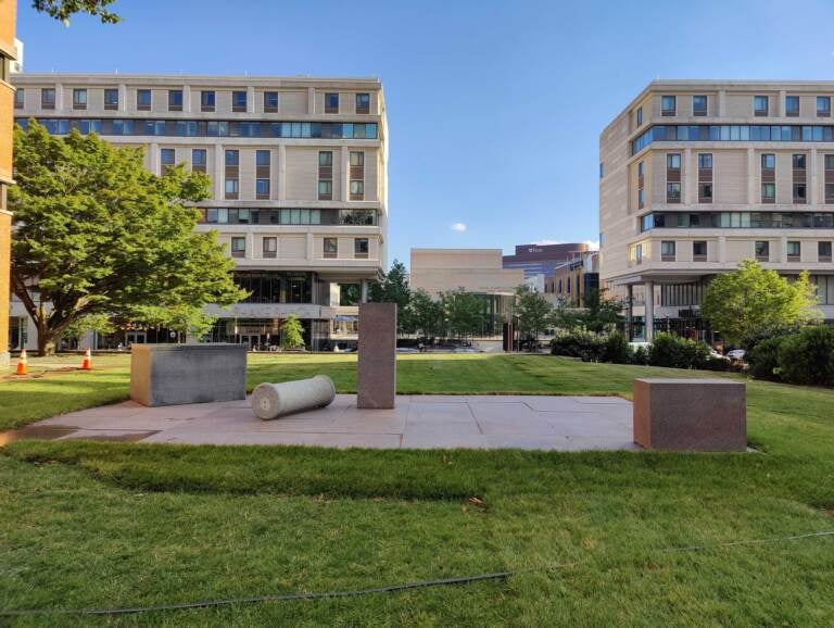 Pillars and pedestals are arranged in an open space, with buildings visible in the background.