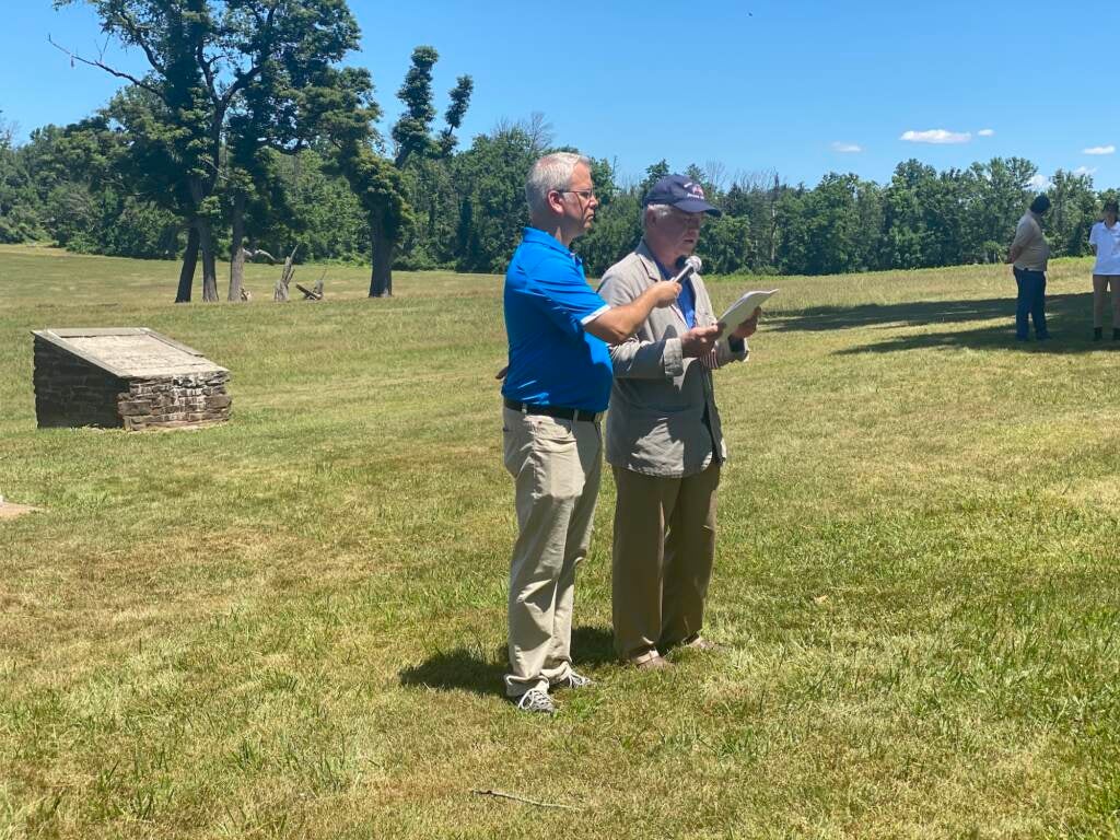 Two men stand in the middle of a field, one reading from a document, the other holding a microphone for him.