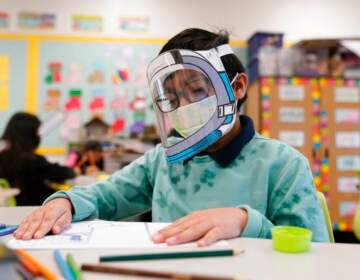 A student sits at a desk wearing a face mask and face shield.