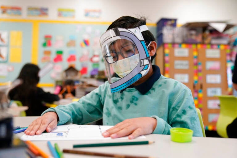 A student sits at a desk wearing a face mask and face shield.