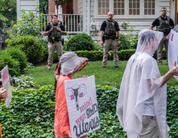 People hold signs and march, with officers standing in the background between protesters and a house.