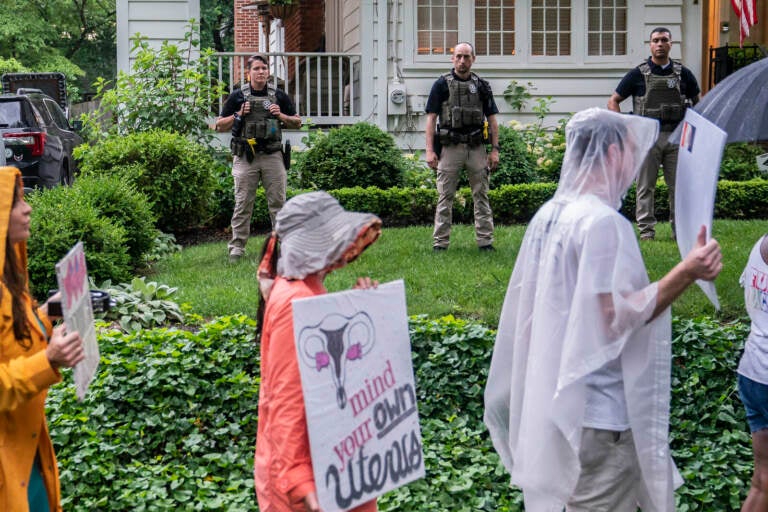 People hold signs and march, with officers standing in the background between protesters and a house.