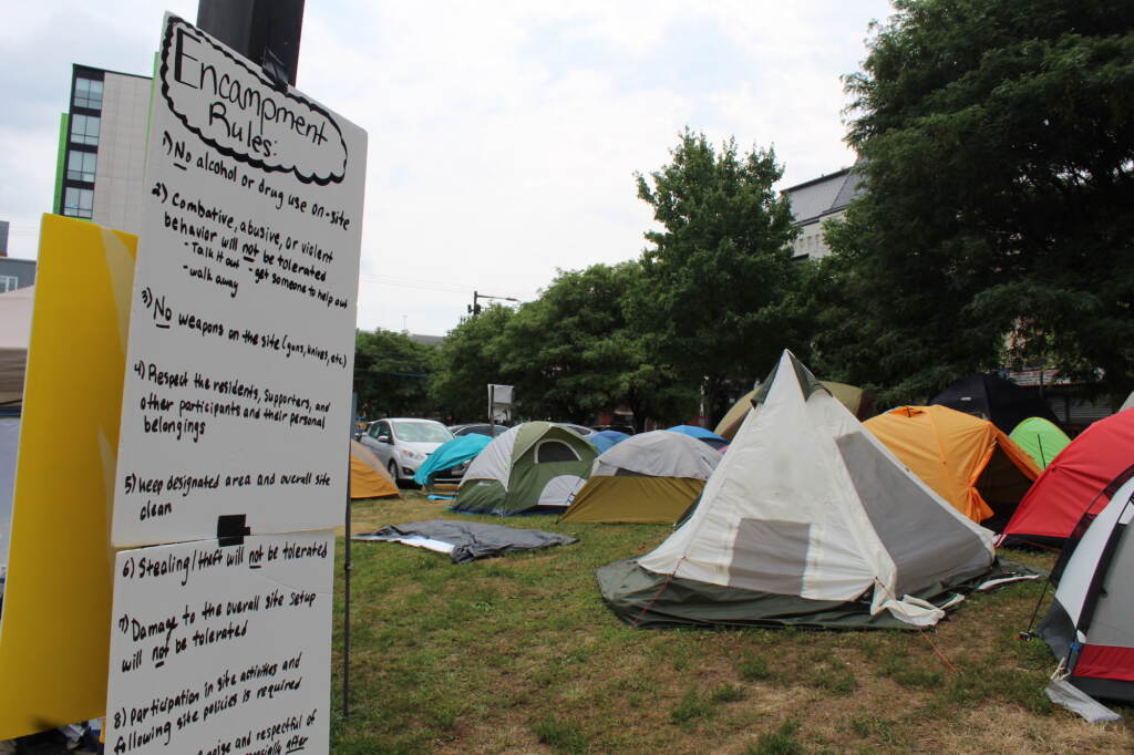 A sign that reads "Encampment rules" is visible in the foreground, with tents visible in the background.