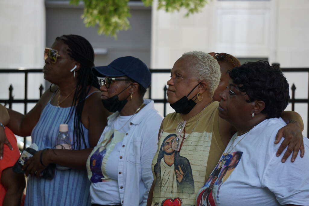 A group of women stand, their arms around one another, looking at something in the distance.