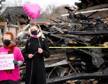 Two people holds a pink balloon in front of a pile of burnt wreckage.